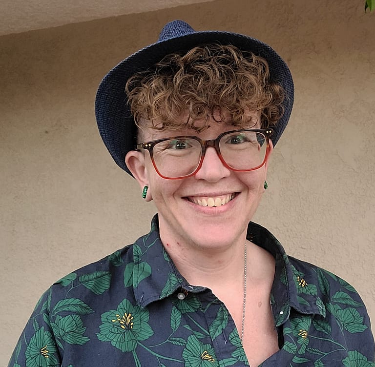 Griffin (they/he) is a white, transmasculine person seen from the shoulders up, posed smiling with a blue hat, curly short hair, glasses, and a casual blue and green floral-print button down shirt.