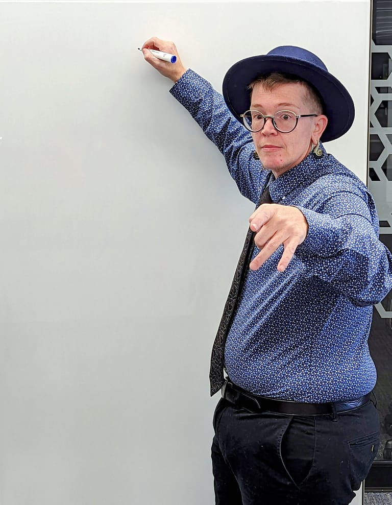 Griffin stands at a whiteboard with one hand raised to write and the other pointing at the camera and audience. He is wearing a dark blue flat-brimmed hat, blue button down dress shirt, gray tie, and gray slacks.